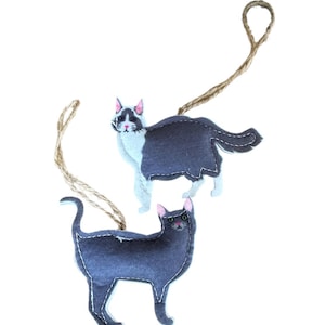 May include: Two felt cat ornaments with twine hangers. One cat is gray and white with pink ears and green eyes. The other cat is gray with pink ears and green eyes.