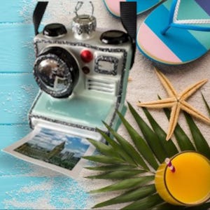 Puede incluir: Adorno decorativo de cámara con una foto, una estrella de mar, un vaso de zumo de naranja y chanclas azules y rosas. La cámara es azul claro con detalles en negro y plata. La foto muestra un edificio con una torre del reloj.
