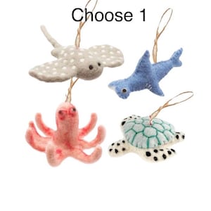 May include: Four felt sea creature ornaments: a stingray, shark, octopus, and turtle. The stingray is gray with white spots, the shark is blue, the octopus is pink, and the turtle is white with a teal shell. Each has a string for hanging.
