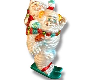 Puede incluir: Adorno navideño vintage que representa a Papá Noel montado en un muñeco de nieve sobre esquís. El adorno tiene una tapa dorada y está decorado con detalles rojos, verdes y azules. El muñeco de nieve y Papá Noel llevan mitones y una guirnalda festiva.