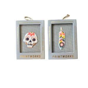 May include: Two small, framed, decorative stickers with a colorful sugar skull and a colorful feather design. The stickers are attached to a white background. The text "PRINTWORKS" is printed on the bottom of each frame.