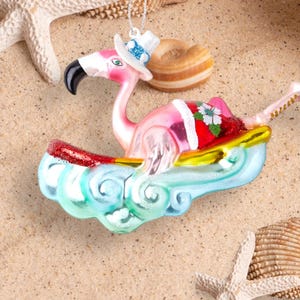 Op de afbeelding: Roze flamingo kerstversiering met een blauwe en witte kerstmanmuts en een surfplank met een rood en geel ontwerp. De versiering is omringd door zand en schelpen.
