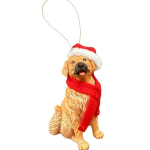 May include: A golden retriever dog ornament wearing a red Santa hat and scarf. The dog is sitting and has its tongue sticking out. A gold string is attached to the top of the ornament for hanging.