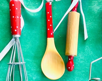 May include: A set of three kitchen utensils: a whisk, a wooden spoon, and a rolling pin. Each has a red handle with white polka dots and is tied with a white ribbon. The whisk is made of silver metal.