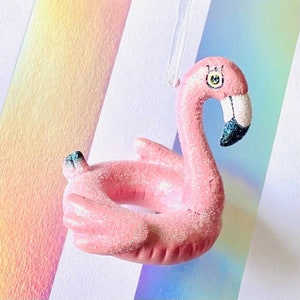 May include: Pink glitter flamingo pool float ornament with a white string for hanging. The flamingo has a black eye with a yellow pupil and a black beak.