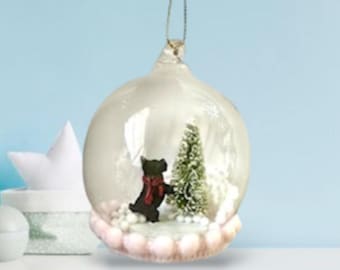 Può includere: Una palla di Natale in vetro trasparente con una statuina di orso nero con una sciarpa rossa all'interno. Un piccolo albero di Natale verde è anche all'interno della palla. La palla ha un cordino dorato per appenderla e una base rosa e bianca.
