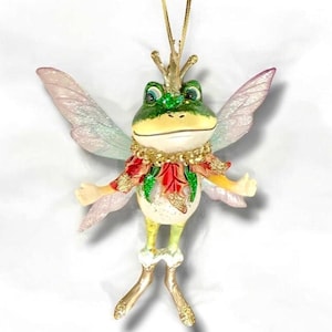 May include: A whimsical frog ornament with fairy wings, a gold crown, and a decorative collar. The frog is primarily green and cream-colored, with red and gold accents on its collar and sleeves. The wings are translucent with a subtle iridescent sheen. A gold string is attached for hanging.