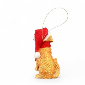 May include: A golden-colored dog ornament wearing a red Santa hat with a white pom-pom. The ornament has a gold string for hanging and is set against a white background. The dog is in a sitting position.