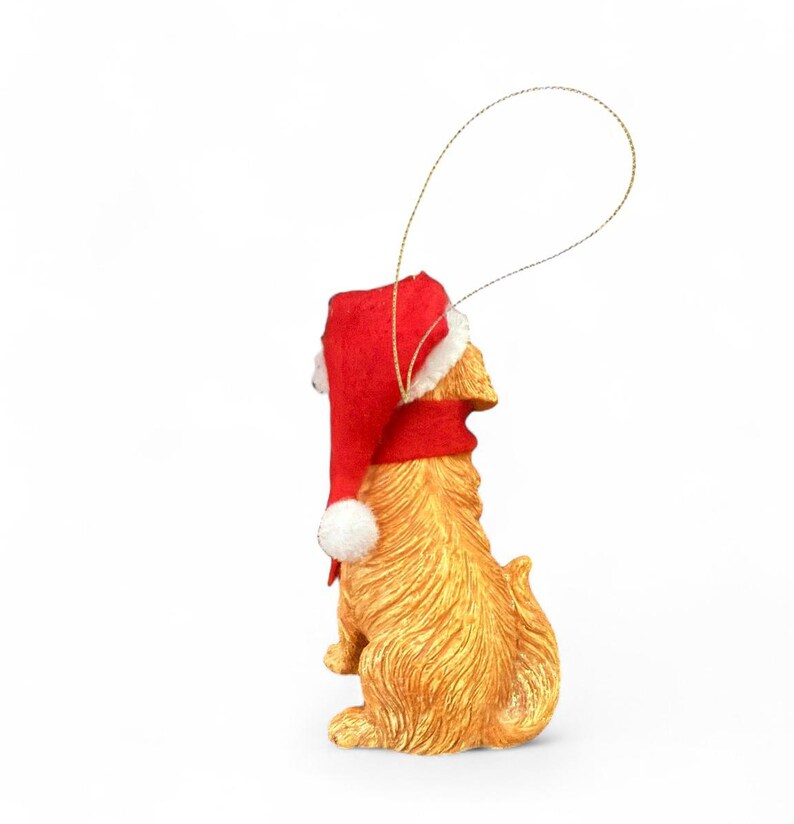 May include: A golden-colored dog ornament wearing a red Santa hat with a white pom-pom. The ornament has a gold string for hanging and is set against a white background. The dog is in a sitting position.