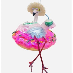 May include: Pink flamingo Christmas ornament with a white straw hat, sunglasses, and a green coconut drink. The flamingo is sitting on a pink and white donut with sprinkles.