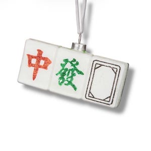May include: A white ornament featuring three Mahjong tiles. The first tile has a red Chinese character, the second a green character, and the third a black outlined rectangle. A silver metal cap and white string are attached for hanging.
