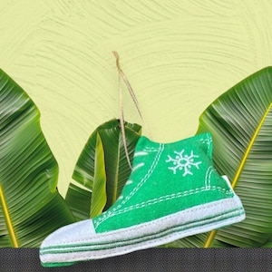 May include: A green and white fabric high-top sneaker ornament with a white snowflake design. The ornament has a white toe cap and white stripes along the sole. A gold string is attached for hanging.