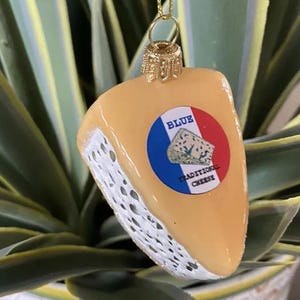 Puede incluir: Un adorno navideño con forma de queso, con una tapa dorada y una cuerda dorada para colgar. El adorno es de color amarillo claro con un borde blanco poroso y una etiqueta circular que dice "BLUE TRADITIONAL CHEESE".