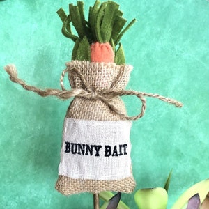 May include: A decorative burlap sack with the text "BUNNY BAIT" on a white label. The sack is tied with twine and topped with a felt carrot and green leaves. The background is a textured teal color.