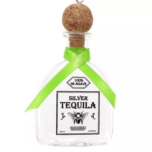 May include: A clear glass bottle ornament shaped like a tequila bottle with a cork stopper and a green ribbon. The label reads "100% DE AGAVE SILVER TEQUILA" with a bee illustration and "HECHO EN MEXICO MADE IN MEXICO 80 PROOF 750 ml".