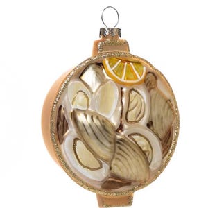 May include: A decorative Christmas ornament shaped like a bowl of clams with a lemon slice. The ornament is primarily beige and gold, with a glittery gold rim. The ornament has a metal loop for hanging.