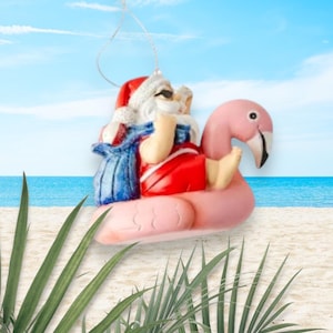 Könnte beinhalten: Ein Weihnachtsbaumschmuck mit dem Weihnachtsmann auf einem rosa aufblasbaren Flamingo. Der Weihnachtsmann trägt einen roten Anzug, einen weißen Bart und eine Weihnachtsmütze. Der Schmuck ist perfekt für ein tropisches Weihnachtsthema.