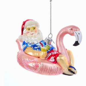 Może przedstawiać: Ozdoba świąteczna przedstawiająca Świętego Mikołaja relaksującego się na różowym dmuchanym flamingu. Święty Mikołaj ma na sobie czerwoną czapkę, żółte okulary przeciwsłoneczne, niebieską hawajską koszulę i czerwone szorty. Trzyma drinka.