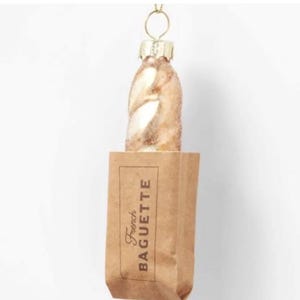 May include: A gold glass ornament shaped like a baguette in a brown paper bag with the text "French Baguette" printed on the front.