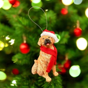 May include: A golden retriever Christmas ornament wearing a Santa hat and a red scarf. The dog is sitting with its tongue sticking out. The ornament is hanging from a silver string against a backdrop of a Christmas tree with red and green ornaments.