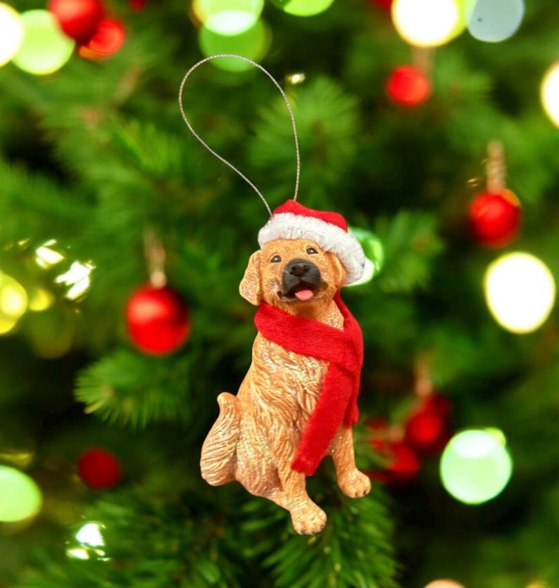 May include: A golden retriever Christmas ornament wearing a Santa hat and a red scarf. The dog is sitting with its tongue sticking out. The ornament is hanging from a silver string against a backdrop of a Christmas tree with red and green ornaments.