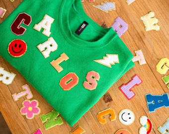 May include: A green sweatshirt with the name "Carlos" spelled out in colorful felt letters. The sweatshirt also has a yellow lightning bolt and a smiley face patch.