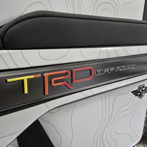 Può includere: Primo piano di un dettaglio interno di un veicolo. L'immagine presenta un rivestimento nero con le lettere "TRD OFF ROAD" in giallo e rosso. Il pannello circostante è bianco con un design a mappa topografica.