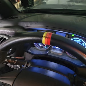 May include: A black leather steering wheel with a colorful stripe design. The steering wheel is in a car interior with a dashboard and gauges visible.