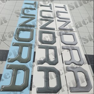May include: Three sets of raised black plastic letters spelling out "TUNDRA" on white and blue backing paper.
