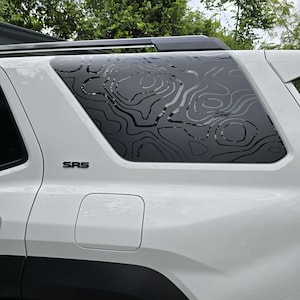 May include: White SUV with a black topographic map design on the rear side window. The window decal is a contour pattern. The letters "SRS" are displayed on the vehicle's body.