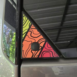 以下が含まれることがあります： カラフルな地形図のデザインが施された、三角形の車の窓トリム。 地図は、黄色、オレンジ、赤の色合いと黒の等高線を使用しています。 黒いボタンがトリムの中央に配置されています。