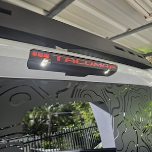 Puede incluir: Una camioneta blanca con una ventana trasera negra y una calcomanía "TACOMA" roja en la parte trasera. La ventana trasera tiene un diseño estampado en blanco y negro.