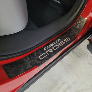 May include: A red car door with a black and gray carbon fiber-style door sill plate. The plate has the words "COROLLA CROSS" in silver lettering.