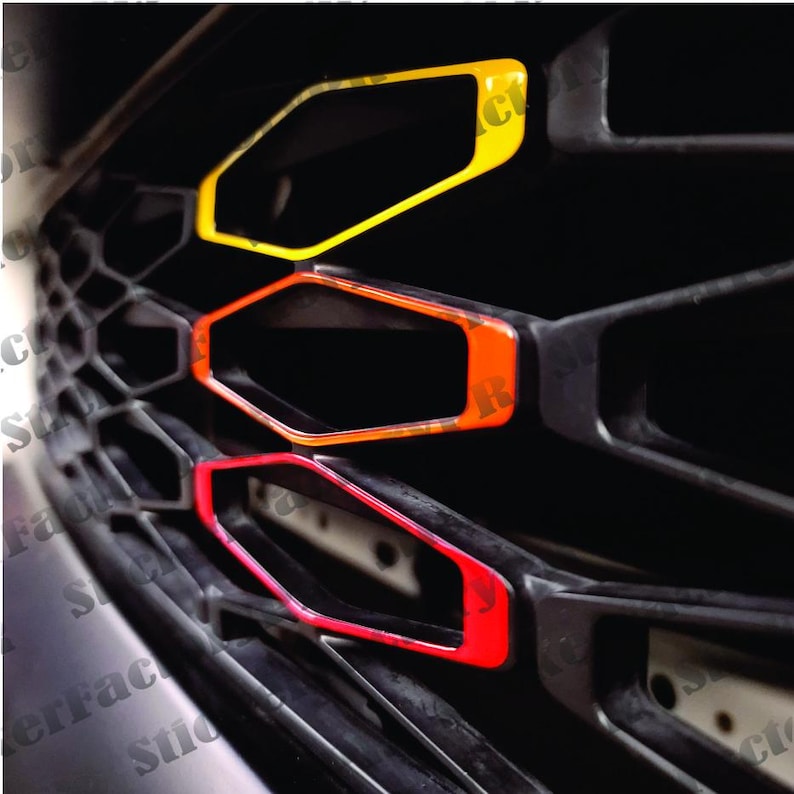 Pu&ograve; includere: Una griglia per auto nera a nido d'ape con tre inserti esagonali individuali di colore giallo, arancione e rosso. Gli inserti sono posizionati verticalmente all'interno della struttura della griglia, evidenziando il motivo geometrico.