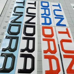 May include: Three sets of raised letters spelling out "Tundra" in black, blue, and red. The letters are on a white background.