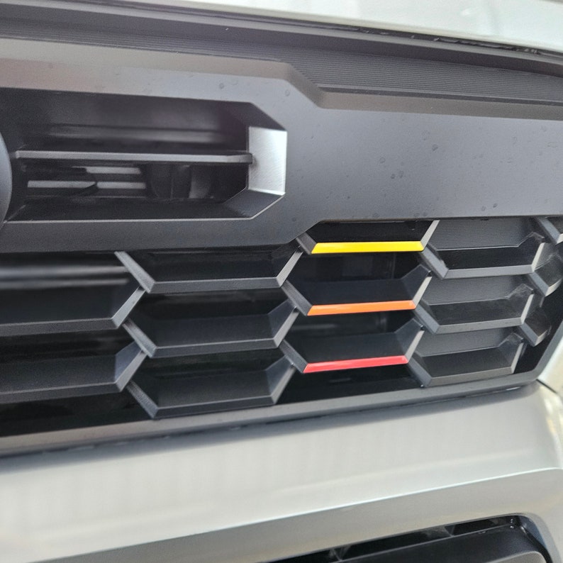K&ouml;nnte beinhalten: Schwarzer LKW-K&uuml;hlergrill mit Wabenmuster und vier horizontalen Streifen in Rot, Orange, Gelb und Gelb.