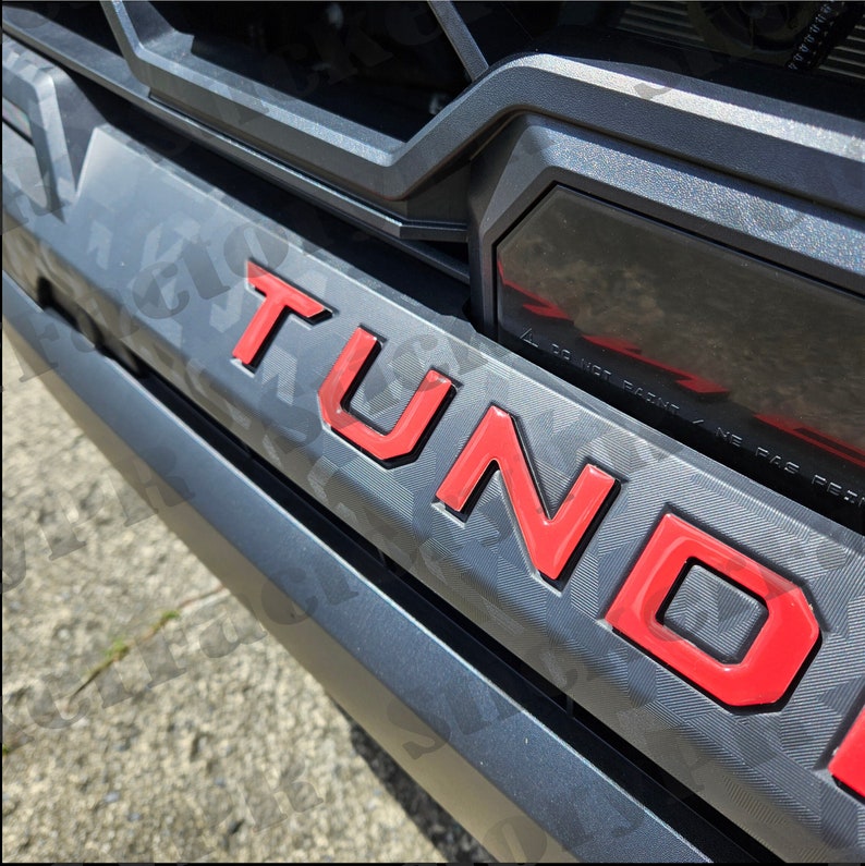 May include: Red "TUNDRA" lettering on a grey truck bumper. The letters are raised and have a glossy finish.