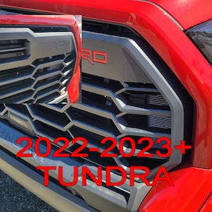 May include: A red 2022-2023+ Toyota Tundra pickup truck with a black grille and "Tundra" lettering. The grille has a honeycomb pattern and a black "TRD" emblem with red lettering.