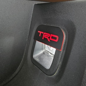 May include: A black plastic panel with a red "TRD" logo. The panel is likely from a car or truck.