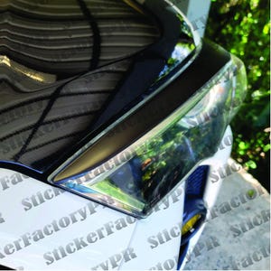 May include: Close-up of a white car's headlight with a black trim. The headlight is partially covered by a black vinyl wrap with a repeating pattern of the words "StickerFactory" and "StickerFactoryPro".