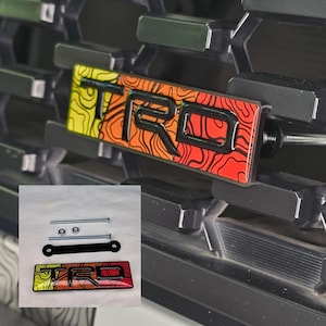 May include: A black metal grille with a colorful "TRO" emblem attached. The emblem has a yellow, orange, and red background with black outlines. The emblem is attached to the grille with a black bracket and screws.
