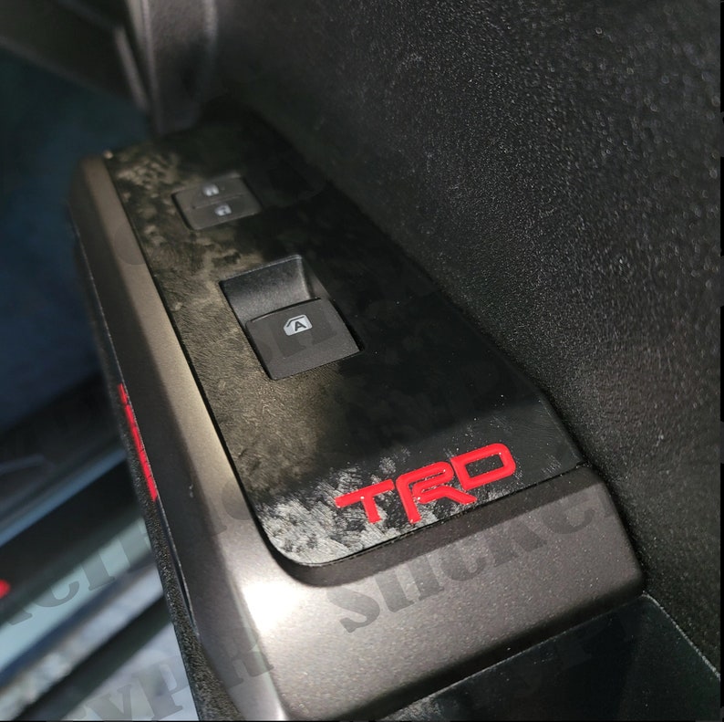 May include: Close-up of a black car door panel with a red "TRD" logo and a window control panel. The panel has a textured, carbon fiber-like finish.