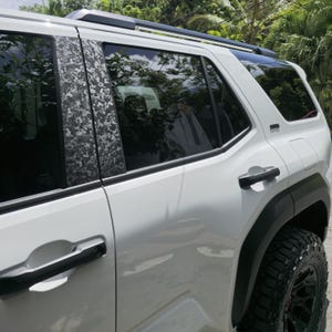 以下が含まれることがあります： 黒いドアハンドル、黒いホイールアーチトリム、ルーフラックを備えた白いSUV。BピラーとCピラーはグレーと黒の迷彩柄で覆われています。車両にはスモークガラスと、黒いリムが付いた大型のオフロードタイヤが装備されています。
