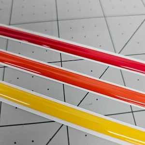 K&ouml;nnte beinhalten: Drei Streifen Klebeband in den Farben Rot, Orange und Gelb. Das Klebeband ist gl&auml;nzend und hat einen wei&szlig;en Rand.