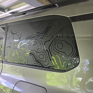 May include: A car window featuring a topographic map design. The black lines form a pattern on the frosted glass, with a green background visible. The car's exterior is a light green colour.