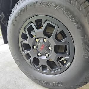 May include: Black tire with a black rim and silver lug nuts. The tire has the words "BFGoodrich" and "Trail-Terrain" written on the side. The rim has the word "TRD" written on it in red.