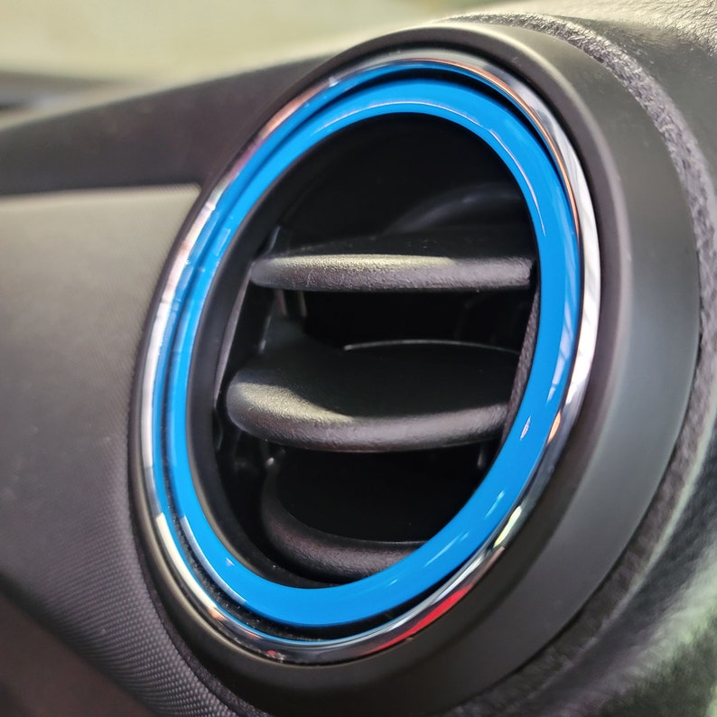 May include: Close-up of a car's air vent with a blue and silver trim ring.