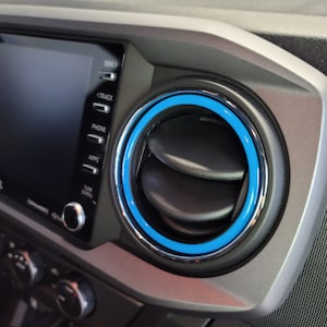 May include: A close-up of a car's dashboard with a blue and silver air vent ring. The vent is circular and has two black flaps. The vent is located next to a touchscreen display with buttons for "Seek", "Track", "Phone", "Apps", "Tune", and "Scroll".