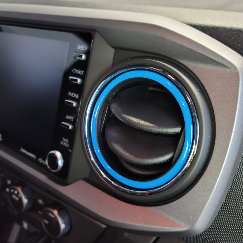 May include: A close-up of a car's dashboard with a blue and silver air vent ring. The vent is circular and has two black flaps. The vent is located next to a touchscreen display with buttons for "Seek", "Track", "Phone", "Apps", "Tune", and "Scroll".