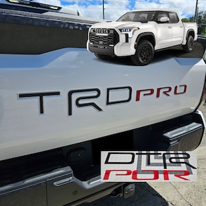 May include: White Toyota pickup truck with a black "TRD PRO" emblem on the tailgate. The truck has a black grille with a chrome Toyota logo and black wheels with red accents.